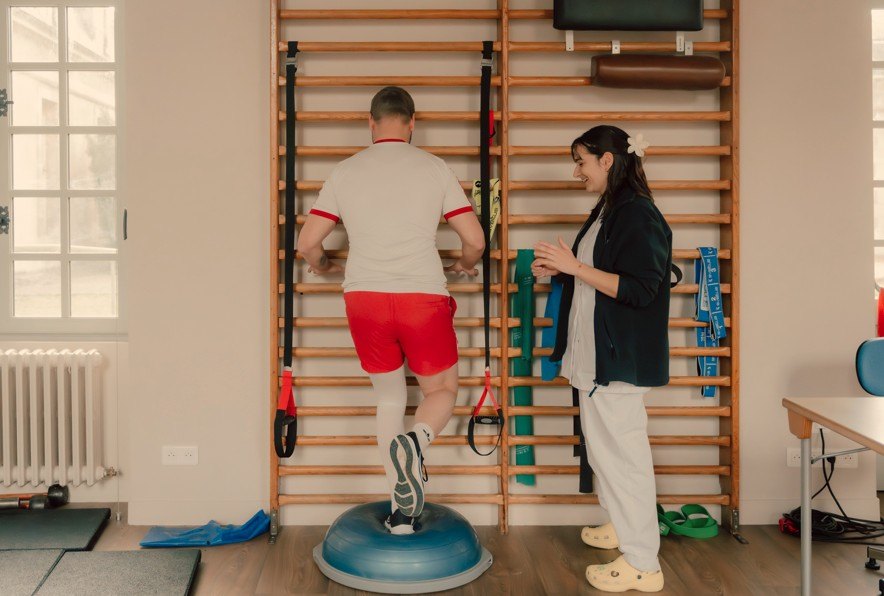Un patient du CRPT réalise un exercice d'équilibre sur plateau instable face aux espaliers, encadré par une soignante